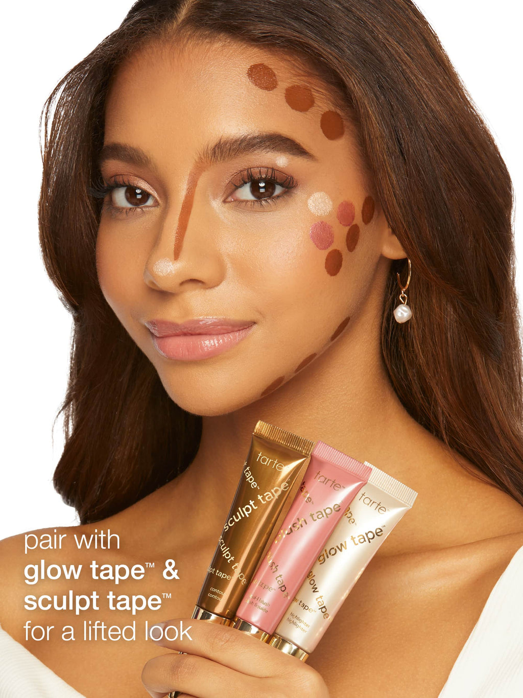 Woman holding three tubes of Tarte Glow Tape and Sculpt Tape with makeup application on her face.