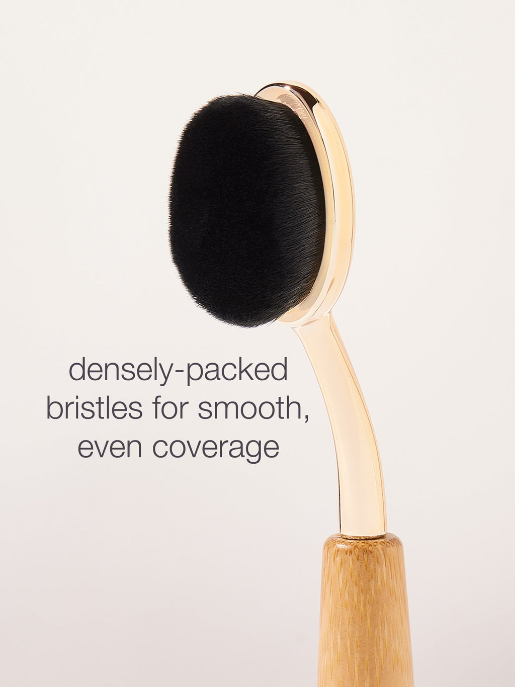 A gold-handled makeup brush with densely-packed black bristles is positioned upright against a plain background. Text reads: densely-packed bristles for smooth, even coverage.