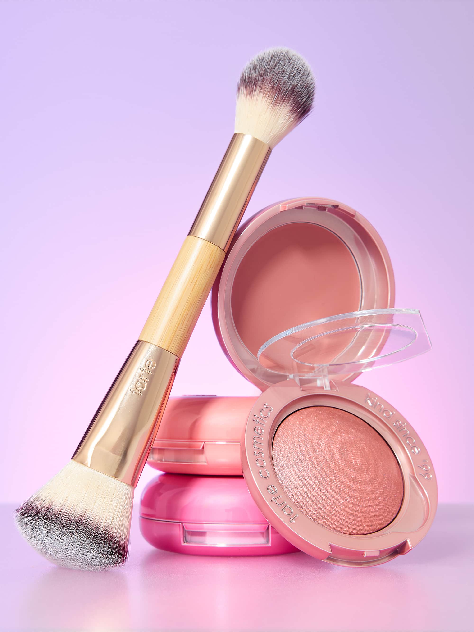 Makeup brush rests across two stacked pink blush compacts; one compact is open, revealing peach powder against a soft purple-pink gradient backdrop. Text: tarte; tarte cosmetics